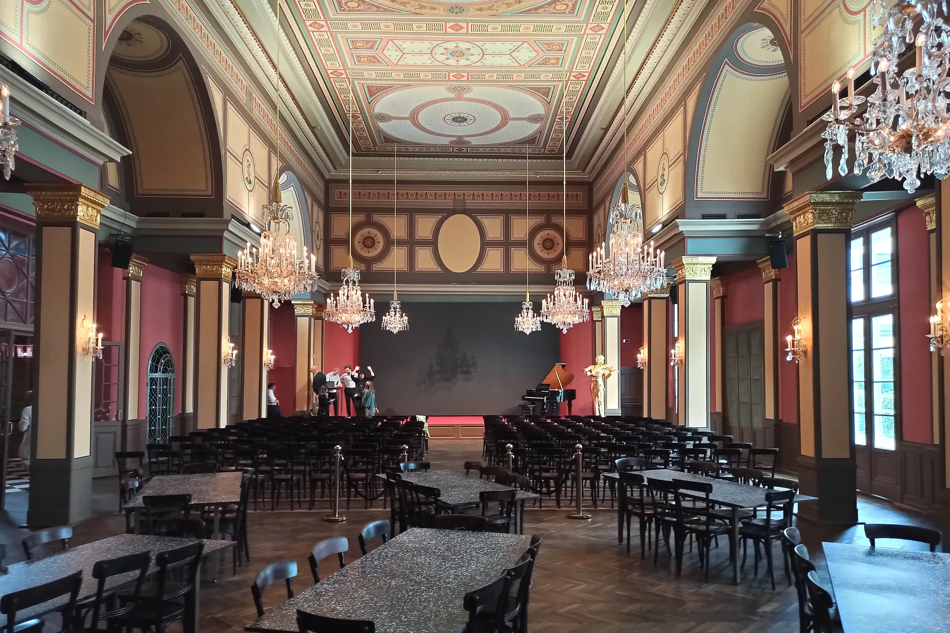The restored concert hall in Vienna's Döbling district: crystal chandeliers hang below a painted ceiling, red walls with gilded capitals frame a stage with a grand piano and a gilded violinist figure, and rows of dark chairs face it