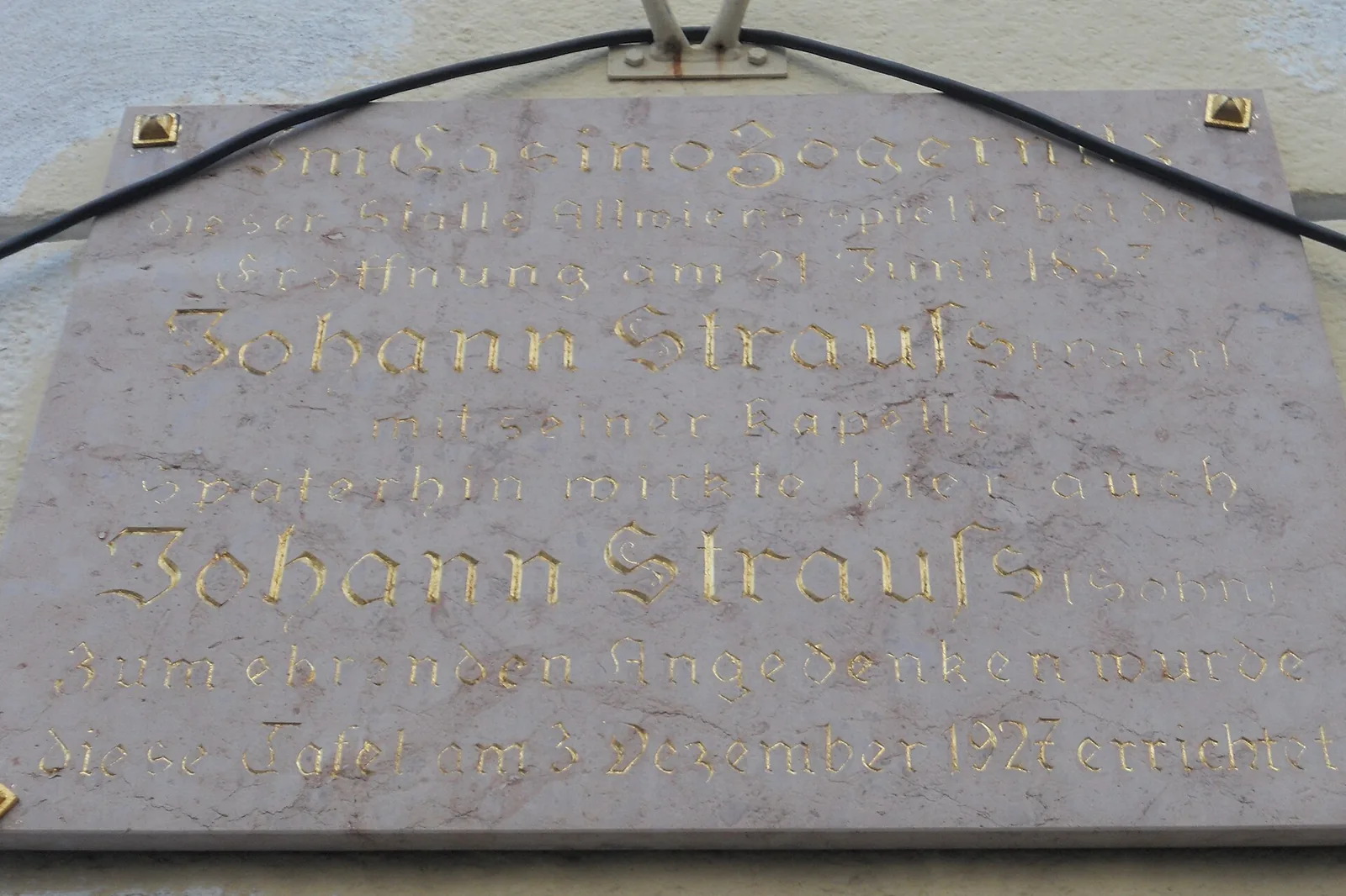 A pale marble plaque on a yellow wall, with gold Gothic-style lettering that names Johann Strauss twice